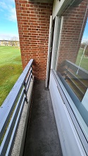 Narrow view of the apartment's balcony with a brick wall on one side and a railing, overlooking a green field.