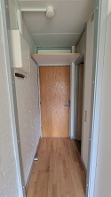 View of the apartment's entrance hallway with a wooden door, light wood flooring, and a small shelf above the door.