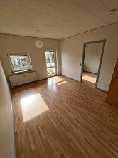 Empty living room with light wooden flooring, white walls, a window, and a door leading to another room.