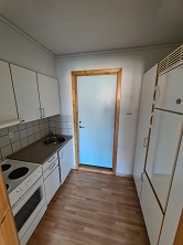 A compact kitchen with white cabinets, a stove, a sink, and a refrigerator, featuring wooden flooring and a light-colored door.