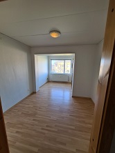 An empty main room with wooden flooring, white walls, and a window providing natural light, viewed from a doorway.