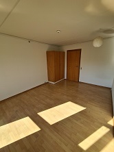 An empty living room with light wooden flooring, white walls, and a corner wardrobe, with sunlight streaming through a window.
