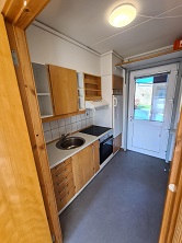 A compact kitchen with light wooden cabinets, a stainless steel sink, an electric hob, an oven, and a refrigerator, with a door leading outside.