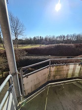 A view from a balcony or outdoor area, overlooking a green landscape with trees under a bright sunny sky.
