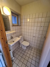 A tiled bathroom with a toilet, a wall-mounted sink, a mirror, and a window, featuring white tiles with dark grout.