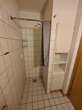 A tiled shower area in a bathroom with a glass shower screen and a white storage cabinet with open shelves.