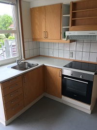 A modern kitchen with light wood cabinets, a stainless steel sink, an induction cooktop, and an oven, with white tiled backsplash.