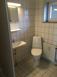 A bathroom with white tiled walls and floor, a toilet, a sink with a mirror above it, and a window.