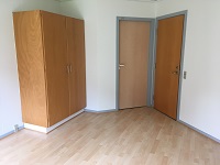 A corner of a room with light wooden flooring, a large wooden wardrobe, and two light-colored doors.