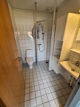 A compact bathroom with white tiled walls and floor, featuring a toilet, shower area with glass screen, and a wall-mounted sink with storage.