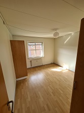 An empty bedroom with light wooden flooring, white walls, a window, and a built-in wardrobe.
