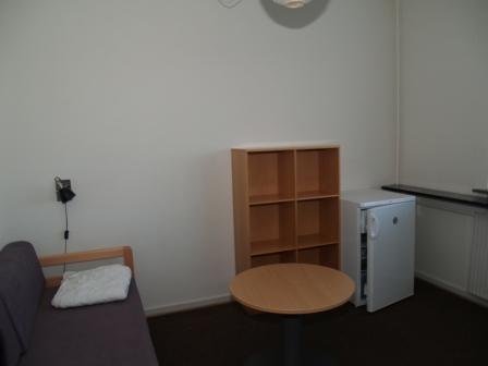 Bedroom or living area showing a bed, wooden shelf, round table, and mini-fridge.