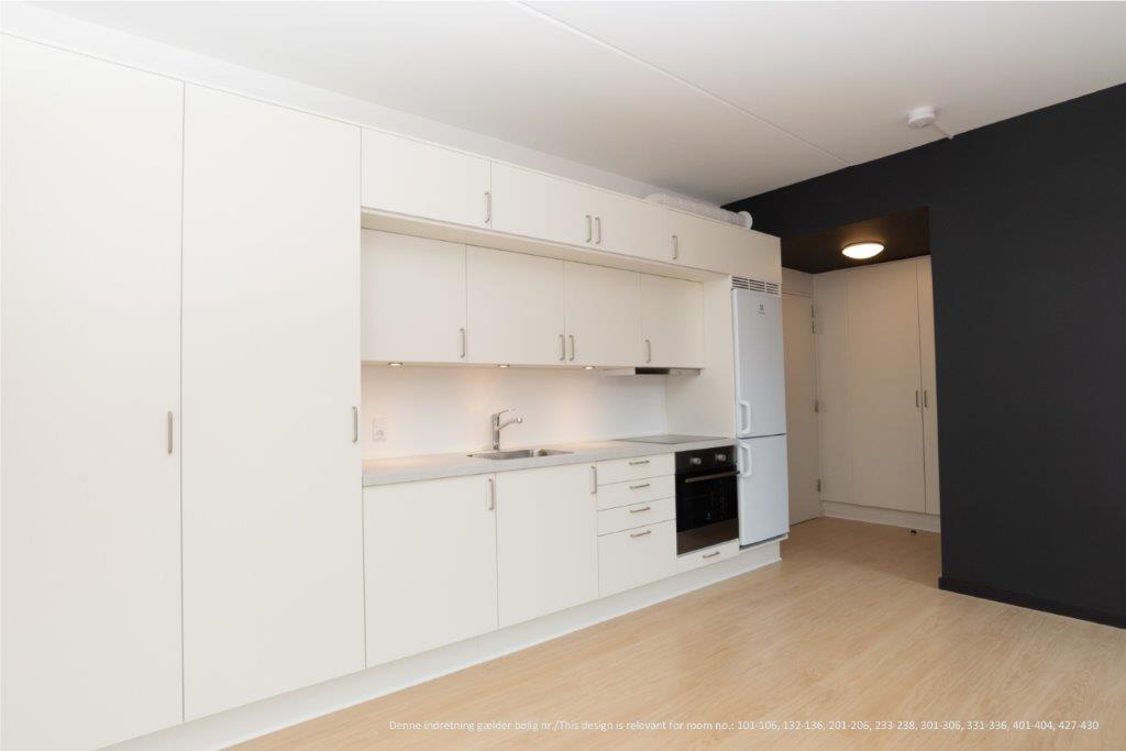 A wide shot of the kitchen area with extensive light-colored cabinetry, integrated appliances, and a light wooden floor, showing a dark accent wall in the background.