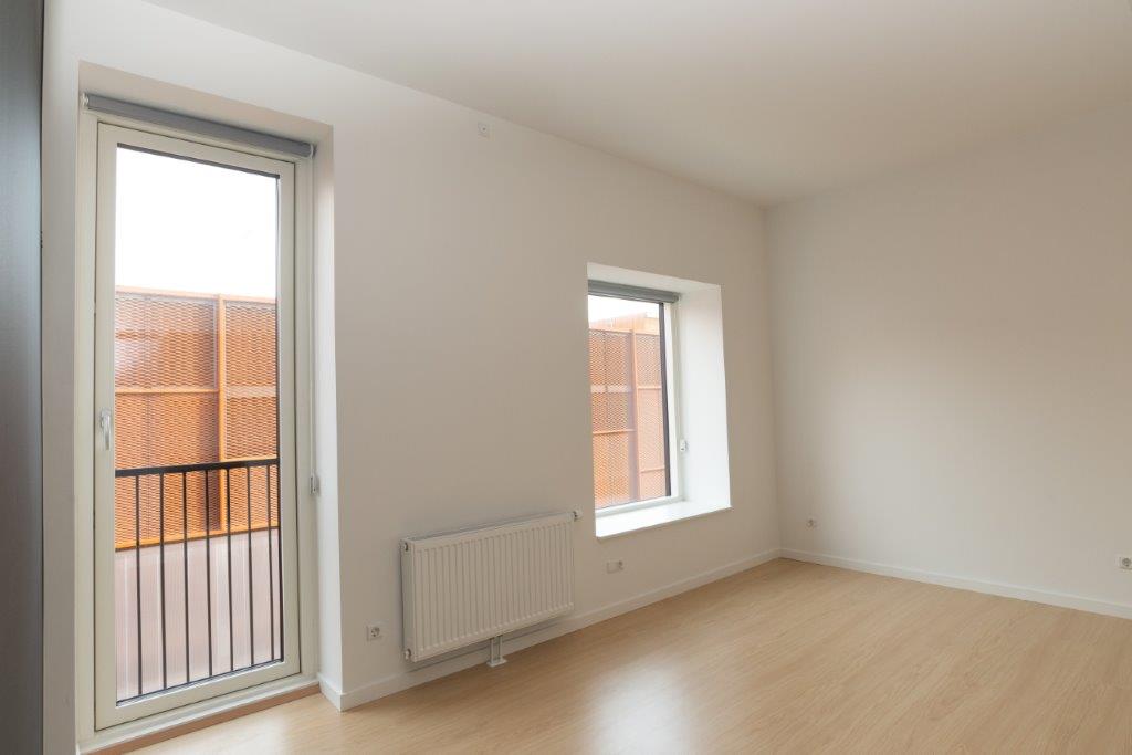 A bright room with light wooden flooring, a large balcony door, and a window, showing a radiator beneath the window.