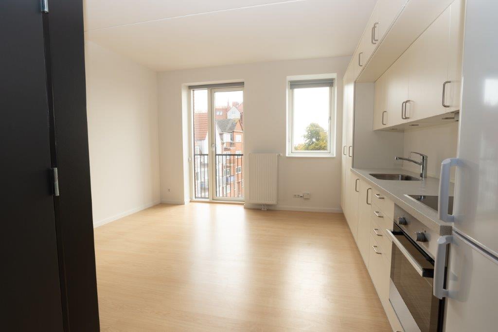 A bright living space with a kitchen area on the right, light wooden flooring, and a balcony door and window offering an exterior view.