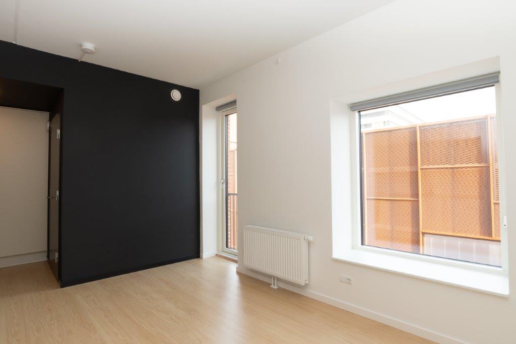 A bright room with light wooden flooring, a dark accent wall, and a balcony door and window providing natural light.