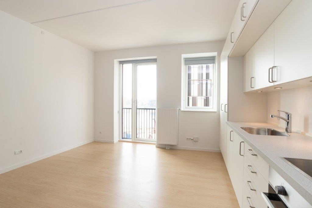 A spacious, empty living area with light wooden flooring, large windows, and a partial view of the kitchen counter.