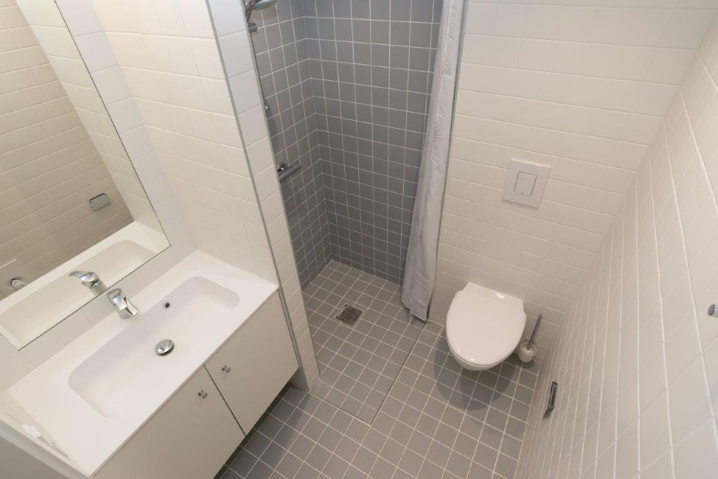 A modern bathroom with white tiled walls, grey tiled floor, a shower area, toilet, and a vanity with a sink and mirror.