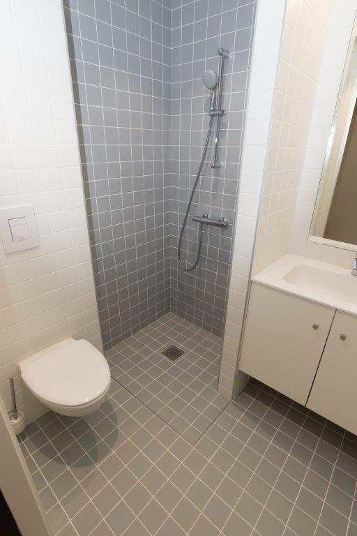 A modern bathroom with grey tiled shower and floor, white tiled walls, and a white toilet and vanity.