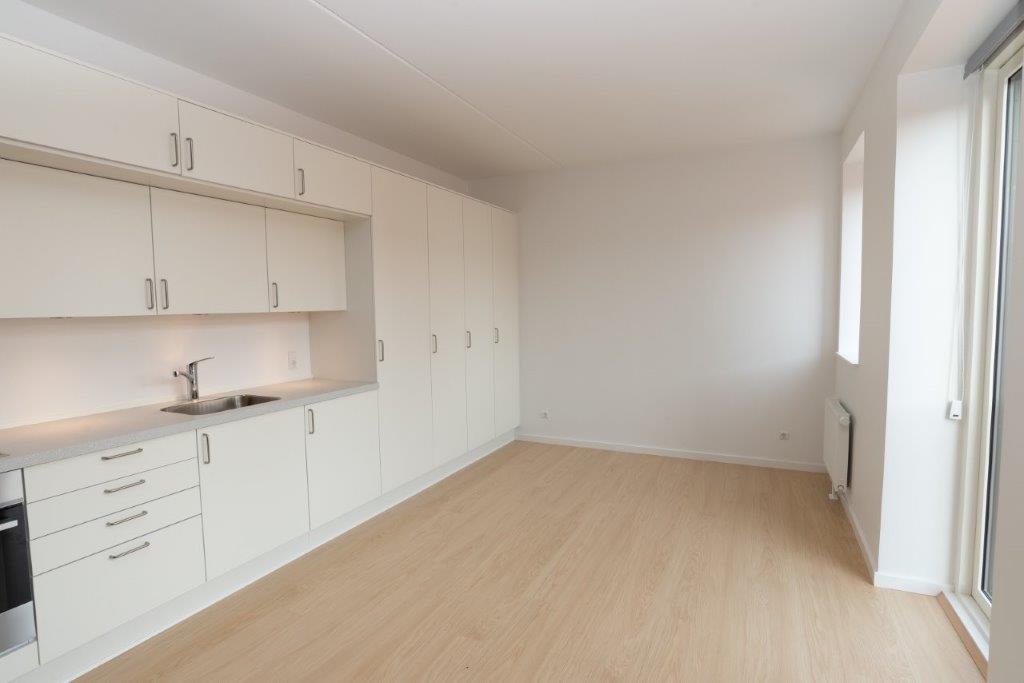 An open-plan living area with light wooden flooring, white walls, and a long white kitchen unit with integrated appliances.