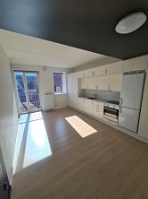 Bright living room and kitchen area with light wood flooring, white cabinets, and a large window with sunlight.