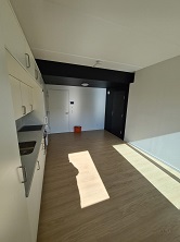 View of the living room and kitchen area looking towards the entrance, featuring light wood flooring and a dark accent ceiling.
