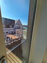 View from a window showing an older building with a pitched roof and a street with water in the distance.