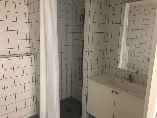 Close-up of a modern bathroom with white tiled walls, a vanity with sink, mirror, and a shower area with a white curtain.
