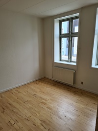 An empty room with light wooden floorboards, white walls, and a large window with a radiator underneath.