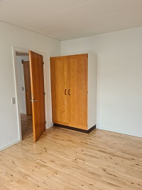 A room with light wooden floorboards, white walls, a wooden wardrobe, and an open door leading to another room.