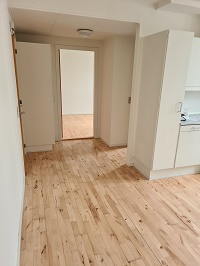 A view from a kitchen area towards an open doorway, showing light wooden floorboards and white walls.