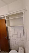 A bathroom view showing white tiled walls, a toilet, and an open white shelf unit above.