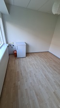 A corner of a room with light wood flooring, a window, and two white appliances, including a refrigerator.