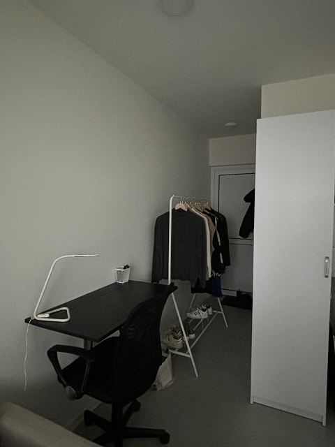 Furnished studio apartment showing a black desk, office chair, and a white clothes rack with garments.