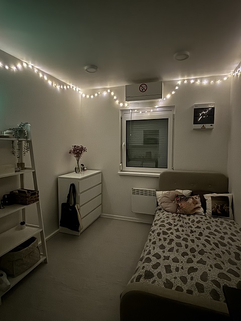 Furnished studio apartment with a bed, white dresser, and a window, decorated with string lights along the wall.