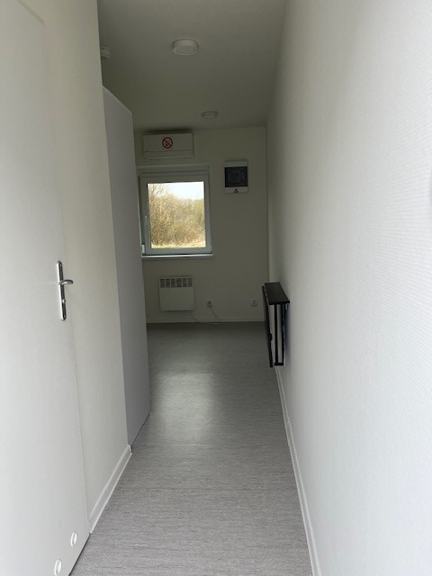 View down a narrow hallway towards a room with a window, showing a radiator and a small shelf.