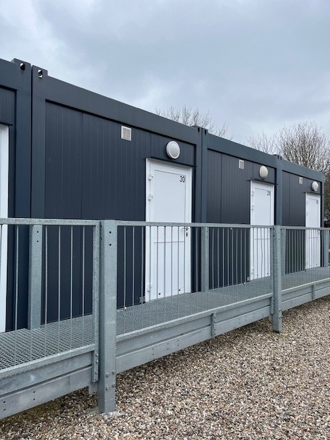Exterior view of modular apartment units with dark grey corrugated facades and white entrance doors, accessed by a metal walkway.