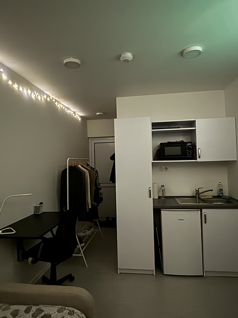 Furnished studio apartment with a desk, office chair, clothes rack, and a compact kitchenette, illuminated by string lights.