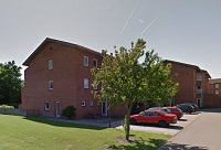 Exterior view of a red brick apartment building with multiple windows, surrounded by a green lawn and a tree, with cars parked in an adjacent lot.