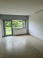An empty, unfurnished room with light-colored walls and a large window and glass door leading to an outdoor area with trees.