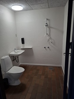A bathroom with white walls, reddish-brown tiled floor, a toilet, a wall-mounted sink, and a shower area.