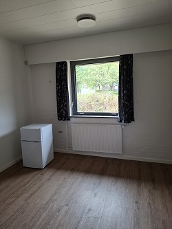 A bright room with light wooden flooring, a window with dark curtains, a white mini-fridge, and a radiator below the window.