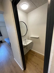 View from a hallway into a small bathroom with light wood-style flooring, a wall-mounted sink, and a round mirror.