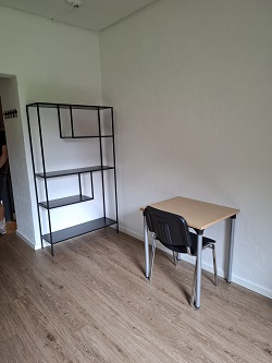 A room with light wood-style flooring, furnished with a small desk and chair, and a modern black metal shelving unit.