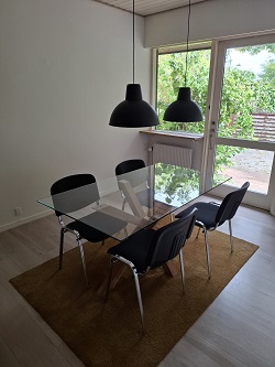 A dining area with a glass-top table, four black chairs, and two black pendant lights, next to a large window.