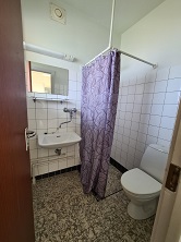 A small bathroom with white tiled walls, a toilet, a sink, a mirror, and a purple shower curtain.