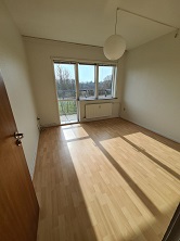 An empty room with light wooden flooring, white walls, a large window, and a door on the left. Sunlight streams through the window.