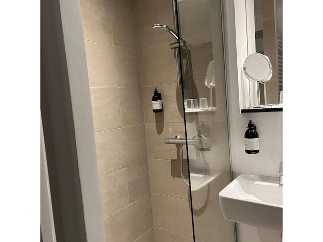 A modern bathroom with beige tiled walls, a glass shower screen, a shower head, a wall-mounted sink, and a round magnifying mirror.