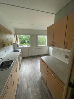 A modern kitchen with light wood cabinets, grey countertops, a stainless steel sink, and a dishwasher, viewed towards two windows.