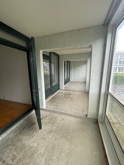 A view from inside a room looking out onto a long, enclosed balcony with large windows and concrete flooring.